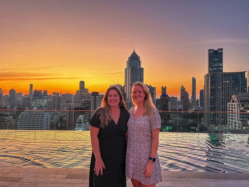 Twee vrouwen staan voor een infinity pool op een rooftop met uitzicht op de skyline van Bangkok tijdens zonsondergang. De lucht kleurt oranje en paars, met wolkenkrabbers op de achtergrond.