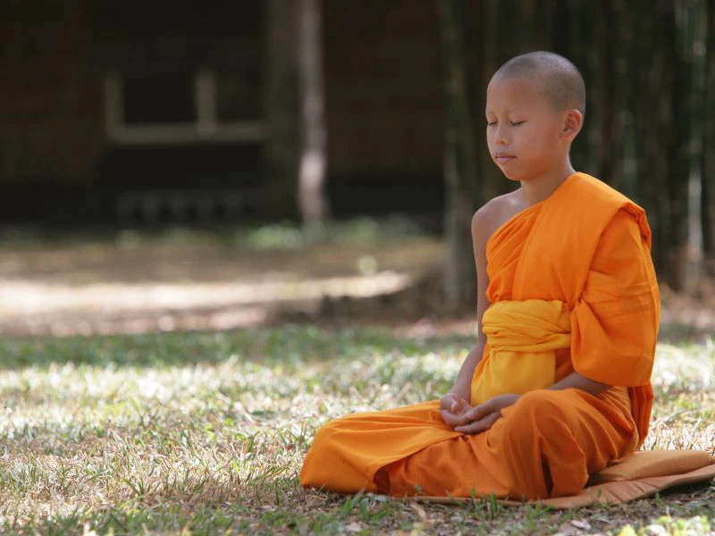 Jonge monnik zit in kleermakerszit op gras, met een oranje gewaad aan.