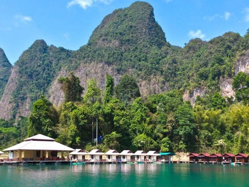 Drijvende bungalows met rieten daken liggen aan een houten steiger op een helderblauw meer, omringd door dichtbegroeide jungle en indrukwekkende, steile kalkstenen rotsen in Khao Sok National Park in Thailand. Op de achtergrond rijzen de groene bergen steil omhoog onder een strakblauwe lucht.