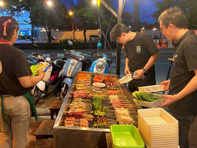 Drie mensen staan bij een straatkraam met verschillende soorten satéstokjes. Op de tafel liggen rijen met groente, vlees, vis en andere ingrediënten op stokjes. Op de achtergrond staan scooters geparkeerd langs de straat.