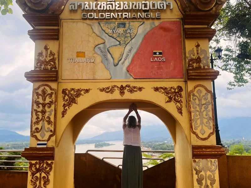 Vrouw staat met geheven armen onder een boog van het Golden Triangle-uitkijkpunt in Noord-Thailand. Boven haar is een reliëfkaart te zien van het drielandenpunt tussen Thailand, Laos en Myanmar. Op de achtergrond stromen de rivieren en zijn de groene heuvels van Laos en Myanmar zichtbaar.