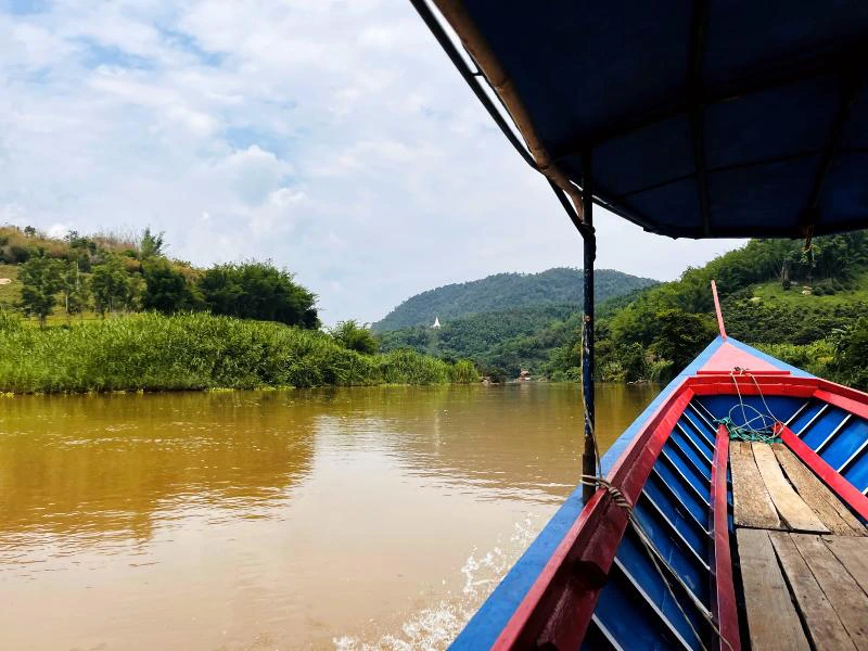 Een houten boot met blauw en rood geverfde zijkanten vaart over een bruine rivier in een groene, heuvelachtige omgeving. Aan de horizon zijn beboste heuvels te zien onder een lichtbewolkte hemel.