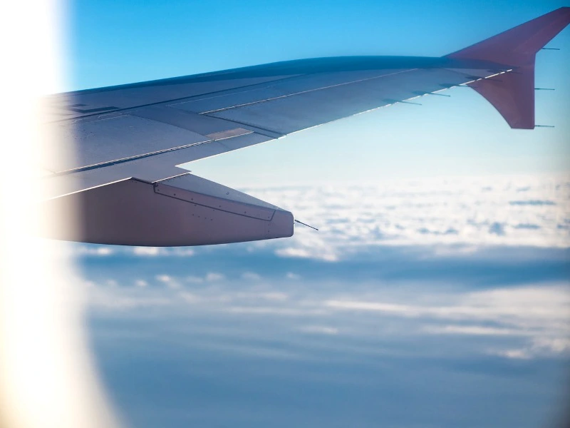 Uitzicht vanuit een vliegtuigraampje op blauwe lucht en wolken, met op de voorgrond de vleugel van het vliegtuig met een rode punt aan het uiteinde.