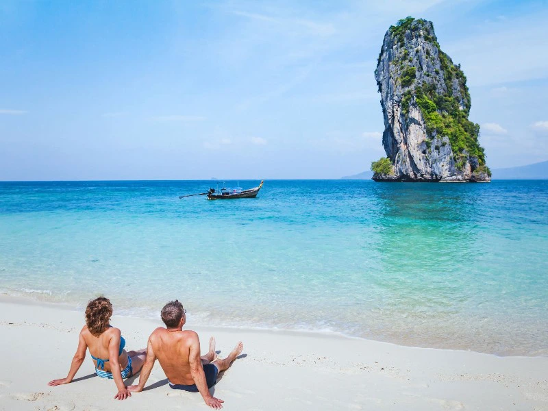 Stel zit op een wit zandstrand met uitzicht op turquoise zee en een steile rotseiland in de verte.
