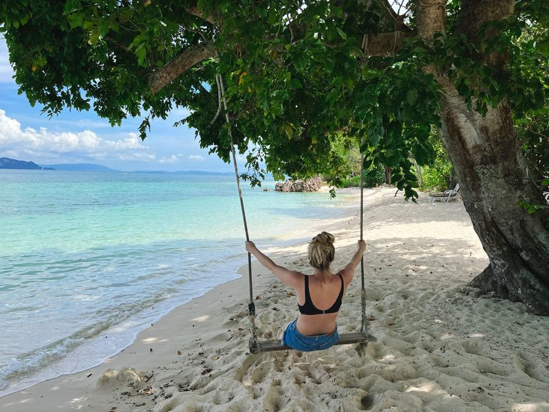 Een vrouw zit op een houten schommel aan een boom, met haar rug naar de camera. Ze hangt boven een zandstrand, vlak bij de zee. De zee is turquoise en kalm, en in de verte zijn eilanden zichtbaar. Er hangt veel groen aan de boom waaronder ze zit.