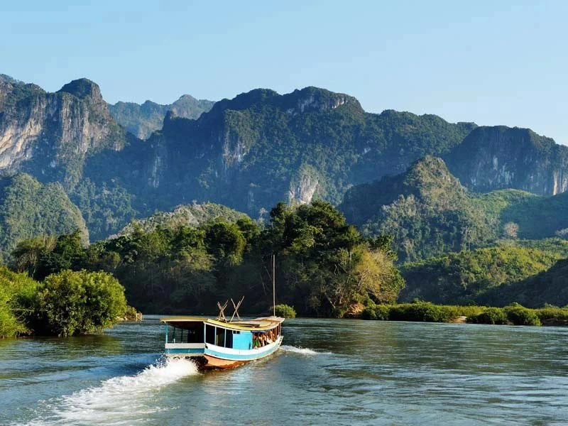Een lang, smal bootje met blauw dak vaart over een brede rivier omringd door groene jungle. Op de achtergrond rijzen steile, ruige kalkstenen bergen op onder een heldere blauwe lucht, ergens in een afgelegen natuurgebied.