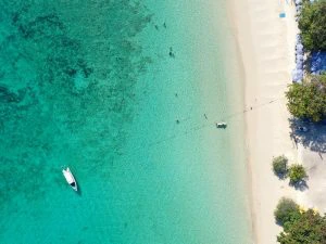 Luchtfoto van een wit zandstrand met helder water. Er liggen mensen in zee, een boot ligt stil voor de kust en aan de rechterkant staan strandparasols en bomen.