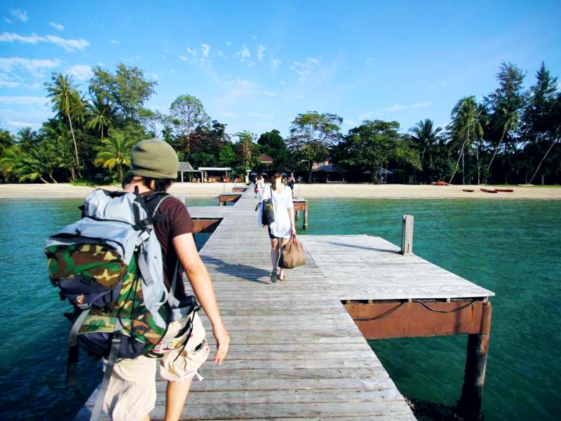 Reizigers lopen met rugzakken en tassen over een houten steiger richting een tropisch strand met palmbomen en helderblauw water.