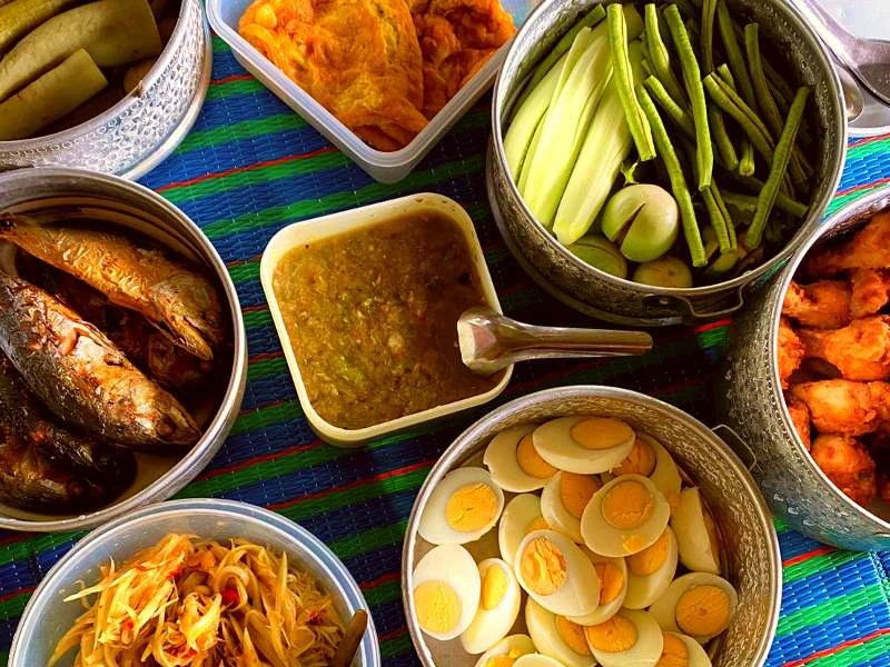 Verschillende Thaise gerechten op een kleed, gekookte eieren, gefrituurde vis, groenten, papajasalade, een groene saus en gefrituurde snacks.