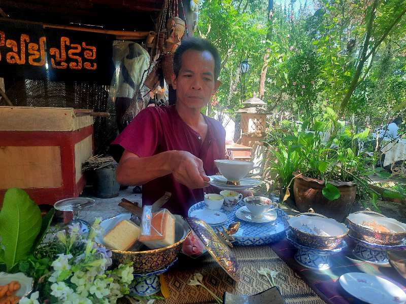 Persoon in paarse kleding schept eten op uit een schaal, zittend achter een tafel met borden, schalen en bloemen, in een groene tuin met beelden en houten decoratie op de achtergrond.