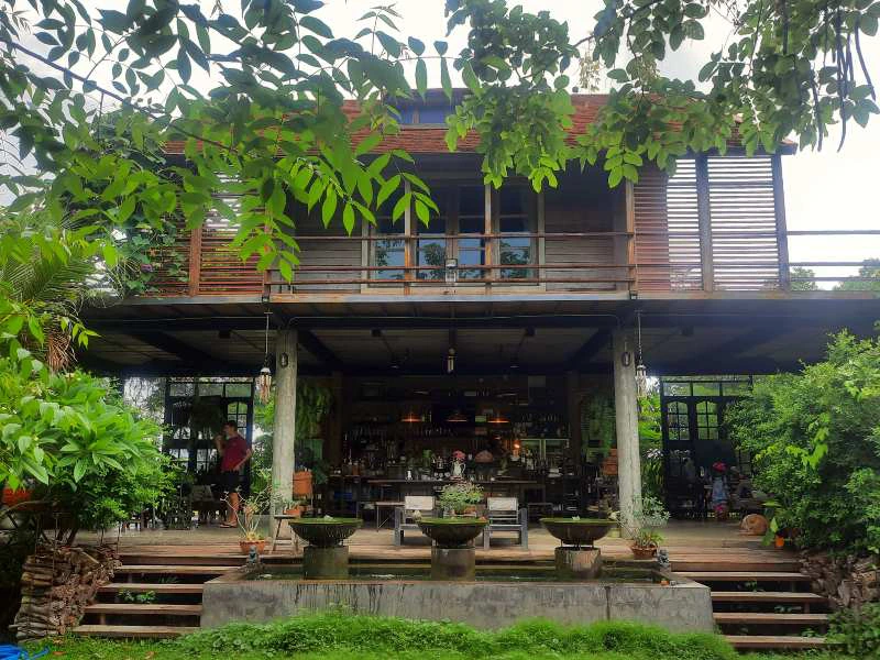 Twee verdiepingen tellend gebouw met houten gevel en grote ramen, omringd door groen. Beneden is een open ruimte met tafels en stoelen, planten in potten en zicht op een bar of restaurantgedeelte.