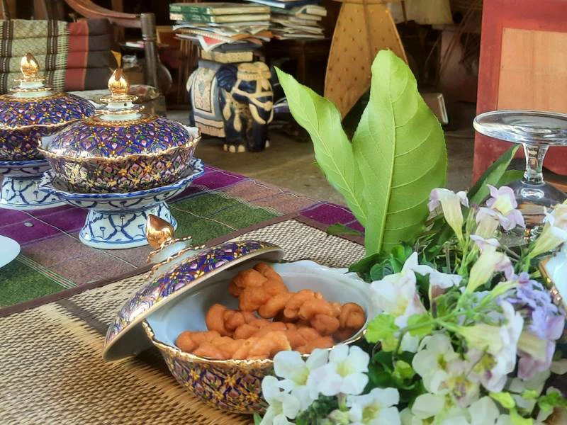 Traditioneel gedekte tafel met rijkversierde Thais porseleinen schaaltjes met deksels, één geopend met oranje snacks erin. Op de voorgrond bloemen en bladeren, op de achtergrond Thaise decoratie met onder andere een keramieken olifant.