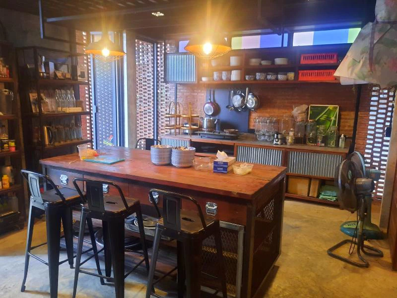 Kleurrijke, industriële keuken met een houten tafel en zwarte barkrukken in het midden. Op de achtergrond open planken met servies, een fornuis met pannen, en decoratieve elementen. Links en rechts metalen roosters voor de ramen, met lichtinval en een staande ventilator op de vloer.