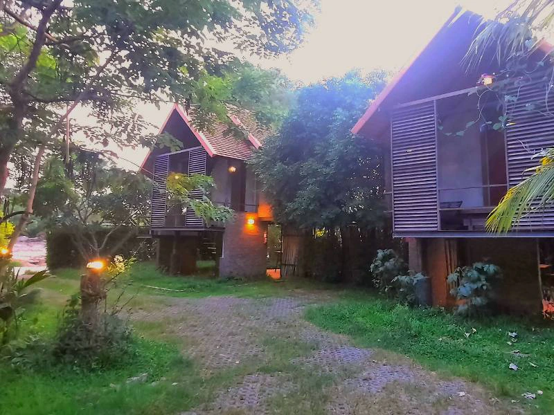 Twee verdiepingen tellend gebouw met houten gevel en grote ramen, omringd door groen. Beneden is een open ruimte met tafels en stoelen, planten in potten