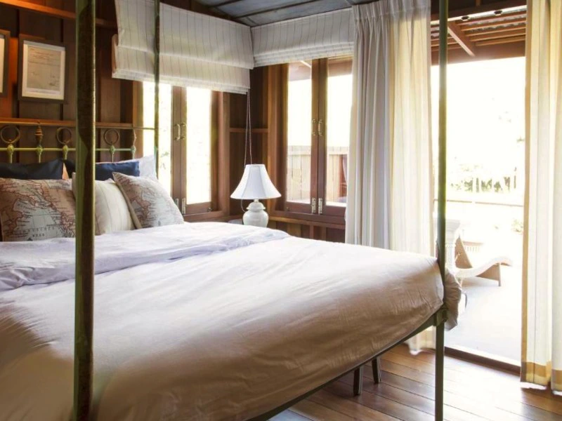 Houten slaapkamer met een groot bed met witte lakens en meerdere kussens. Naast het bed staat een witte lamp op een nachtkastje. De kamer heeft houten wanden, ramen met witte gordijnen en een schuifdeur naar een veranda met uitzicht naar buiten.