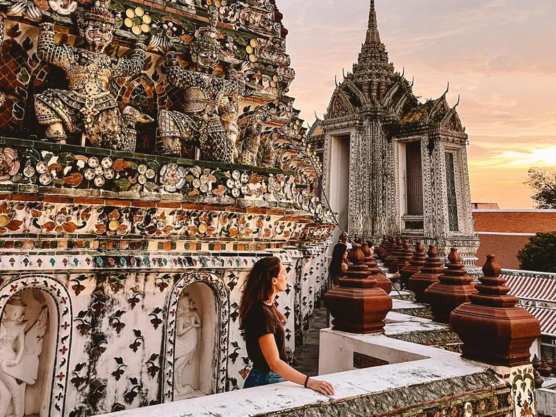 Een vrouw kijkt uit over een rijkversierde tempel in Bangkok met kleurrijke mozaïeken en beelden van wachters. Op de achtergrond is een tweede tempelgebouw te zien met een spitse toren, terwijl de lucht warm kleurt door de ondergaande zon.