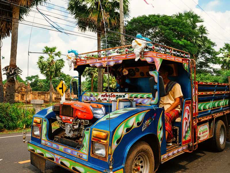 Kleurrijke Thaise vrachtwagen met sierlijke beschildering rijdt over een weg omringd door palmbomen; een man in traditionele kleding zit aan het stuur.
