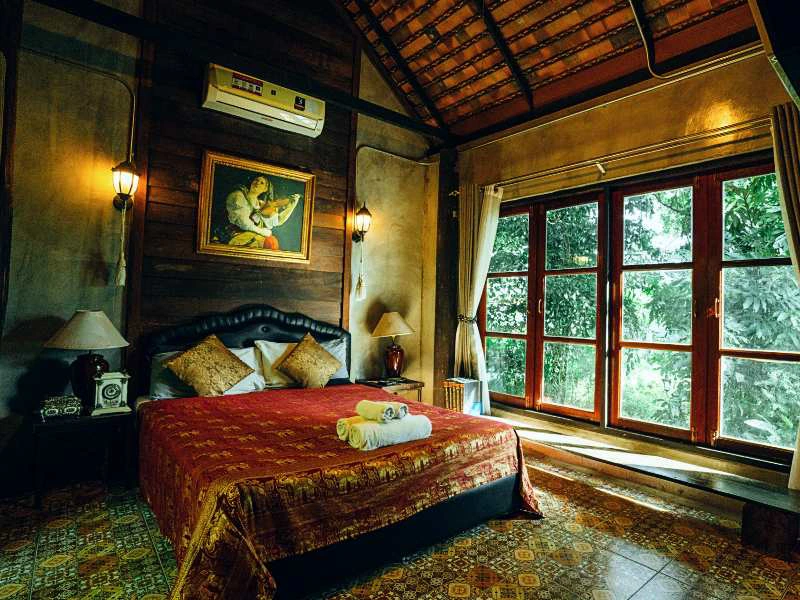 Knusse slaapkamer met groot bed, rood dekbed en sierkussens, twee nachtkastjes met lampen, een schilderij aan de muur en een groot raam met uitzicht op groene bomen.