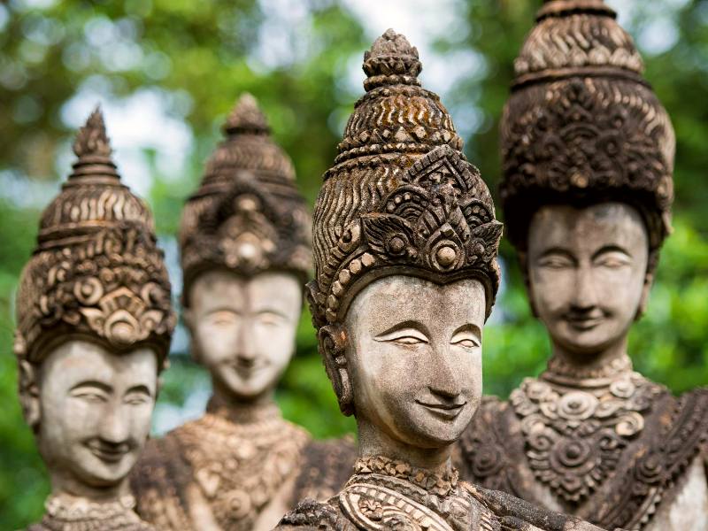 Vier gedetailleerde stenen beelden met sierlijke hoofdbedekking en vriendelijke gezichtsuitdrukking, geplaatst in een groene buitenomgeving.