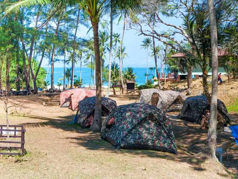 Kleurrijke tenten verspreid over een kampeerplek onder palmbomen, met uitzicht op zee en een tropisch strand op de achtergrond.