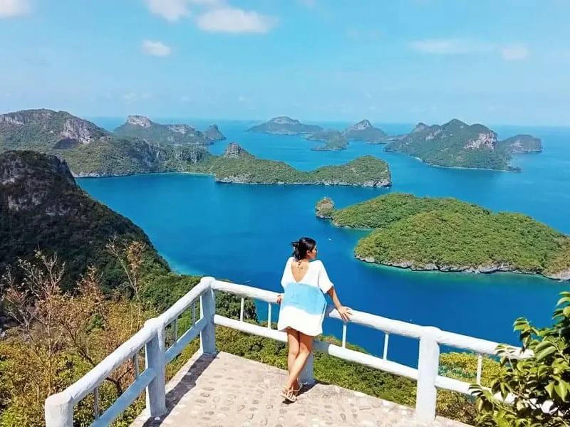 Vrouw op een uitkijkpunt met wit hekwerk, kijkend over een helderblauwe zee met groene eilanden en rotsformaties onder een zonnige hemel.