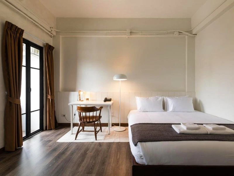 Minimalistische hotelkamer met een tweepersoonsbed, wit bureau met houten stoel, staande lamp en opgevouwen handdoeken op het bed, naast een glazen deur met bruine gordijnen.