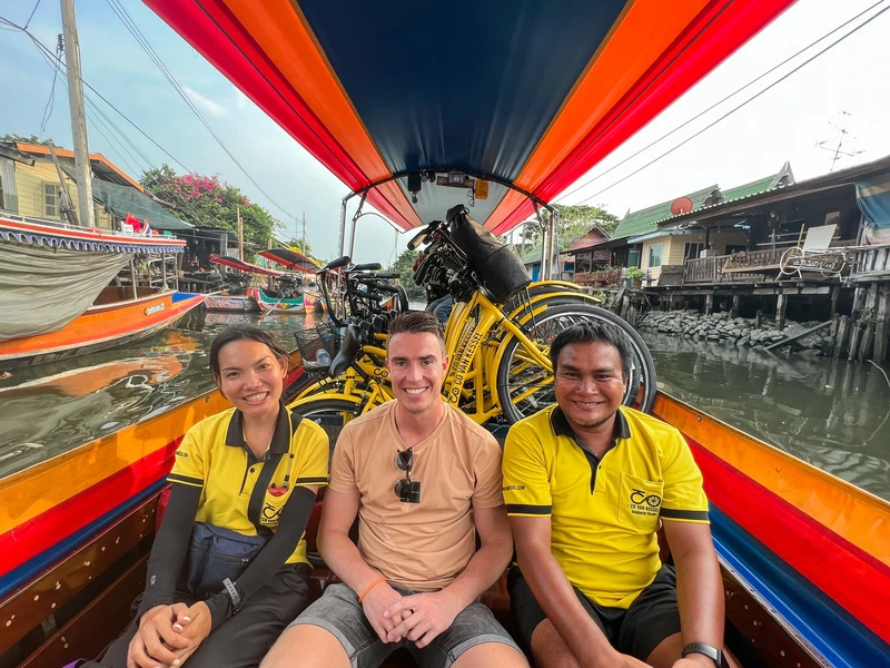 Drie mensen glimlachen in een kleurrijke longtailboot vol met fietsen, varend door een kanaal langs houten huizen in Thailand.