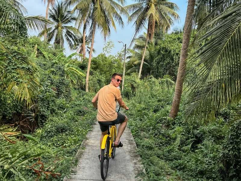 Man fietst op een smal pad door tropisch groen met palmbomen, kijkt glimlachend achterom.