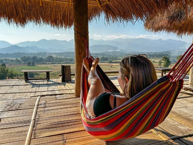 Vrouw ligt in een gekleurde hangmat onder een rieten dak en kijkt uit over groene velden en bergen in de verte.