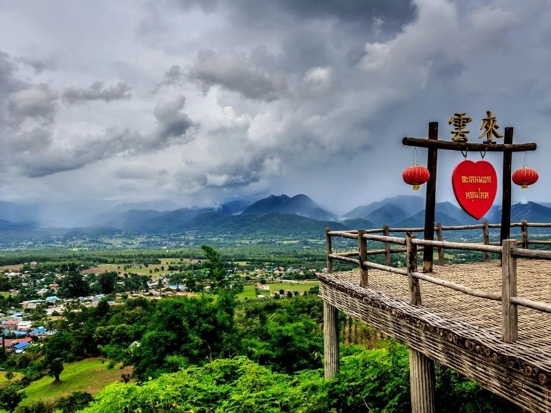 Houten uitkijkplatform met rood hartbord en Chinese lantaarns, uitkijkend over een groen dal met bergen en dreigende wolken.