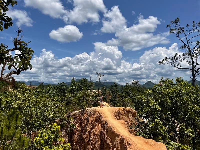 Een persoon loopt op een smal zandstenen pad dat boven de bomen uitsteekt, met op de achtergrond heuvels en een blauwe lucht vol wolken.