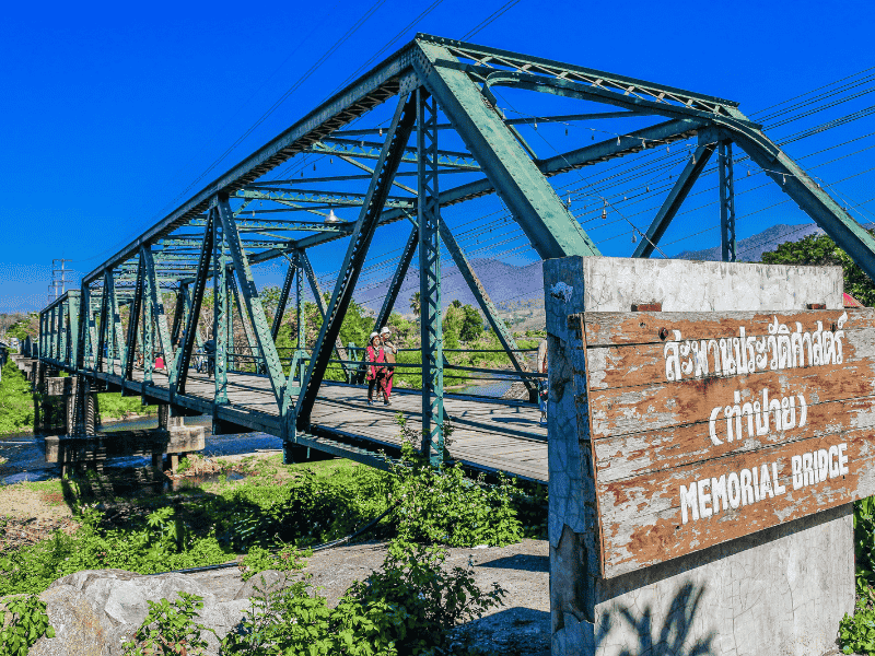 Groene metalen brug met houten vloer over een rustige rivier, omringd door groene begroeiing; een koppel in rode kleding loopt over de brug. Op de voorgrond een houten bord met Thaise letters en de tekst "Memorial Bridge" in het Engels, onder een strakblauwe lucht.