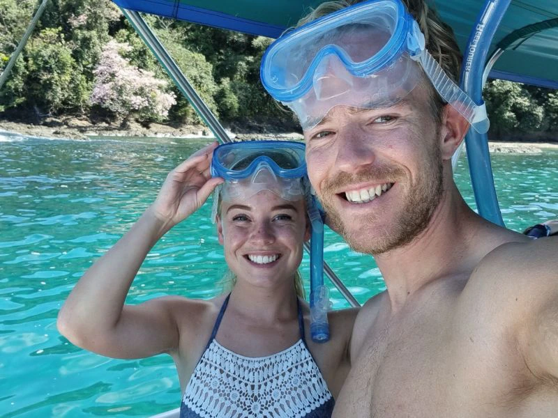 Twee reizigers met snorkelmaskers poseren lachend op een boot boven helderblauw tropisch water.