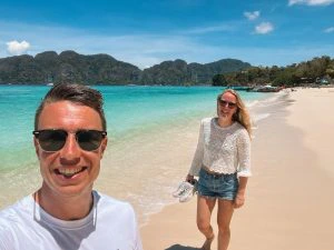 Twee reizigers wandelen lachend langs een tropisch zandstrand met turquoise zee en groene eilanden op de achtergrond. Blauwe lucht