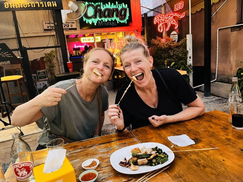 Twee vrouwen zitten lachend aan een houten tafel op een avondmarkt en eten spiesjes. Op tafel staan dipsausjes en een bord met streetfood. Op de achtergrond kleurrijke neonreclames.