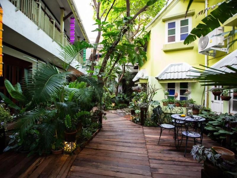 Houten looppad tussen gebouwen met veel planten en bomen, links een wit gebouw met balkon en rechts een lichtgroen huis met witte luifels en een zwart metalen tuinsetje.