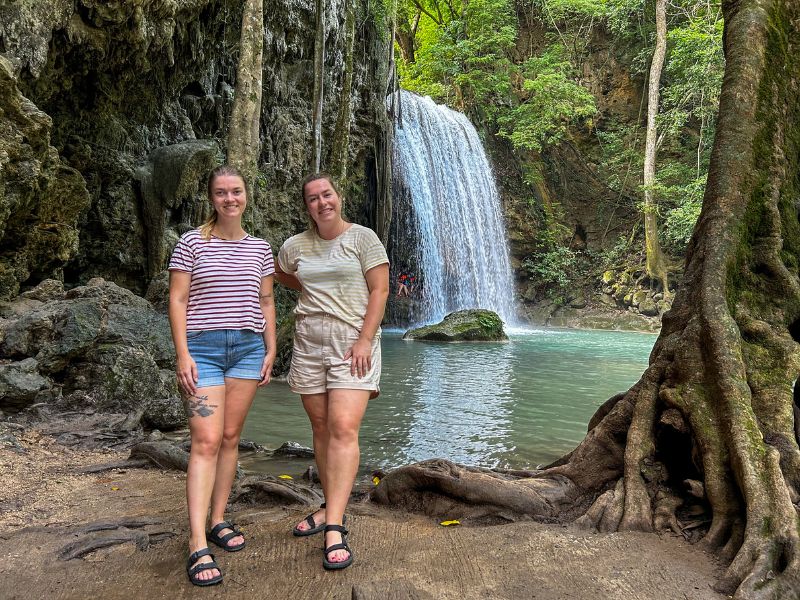 Twee vrouwelijke reizigers poseren voor een riviertje met een waterval, omringd door een rotswand en junglebegroeiing.