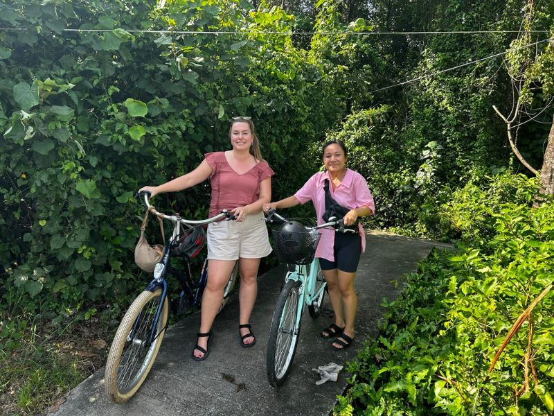 Twee vrouwelijke reizigers met fietsen op een smal pad omringd door dichte groene begroeiing.