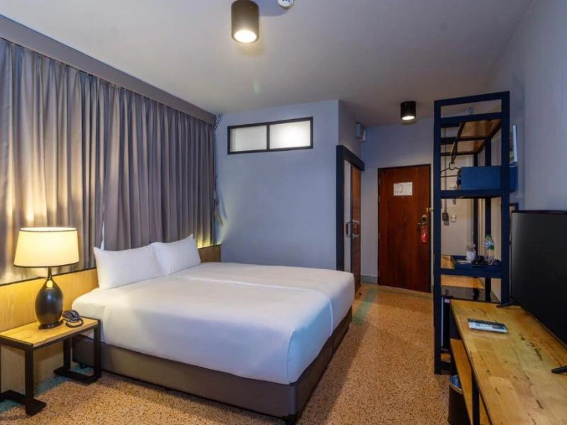 Je ziet een moderne, eenvoudige hotelkamer met een tweepersoonsbed, houten bureau, open kast en tv. De kamer heeft een rustige uitstraling met grijze gordijnen en zachte verlichting.