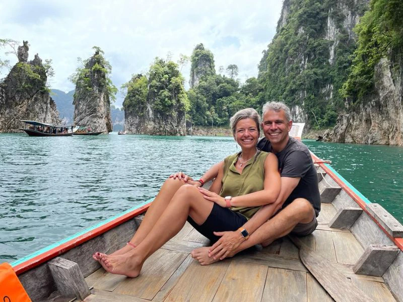 Twee reizigers zitten samen op de punt van een longtailboot op het water tussen kalksteenrotsen.