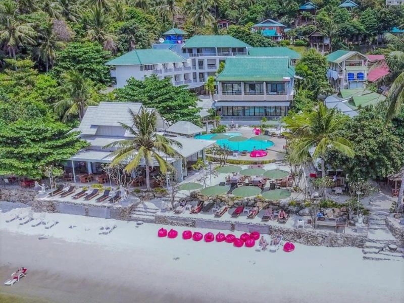 Vogelvluchtfoto van een strandhotel omringd door tropisch groen. Voor het hotel liggen felroze zitzakken op het witte zandstrand, met daarachter een zwembad, ligbedden en parasols. Het complex bestaat uit meerdere gebouwen met groene daken en ligt direct aan zee.