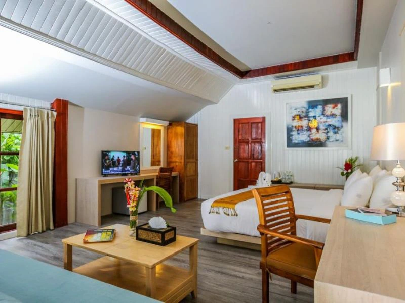 Ruime hotelkamer met een houten vloer en lichte muren. De kamer is ingericht met een groot tweepersoonsbed, houten meubels, een zitgedeelte met salontafel en een bureau met televisie. Er hangen kleurrijke kunstwerken aan de muur en op tafel staan bloemen. Een schuifdeur biedt uitzicht op tropisch groen buiten.