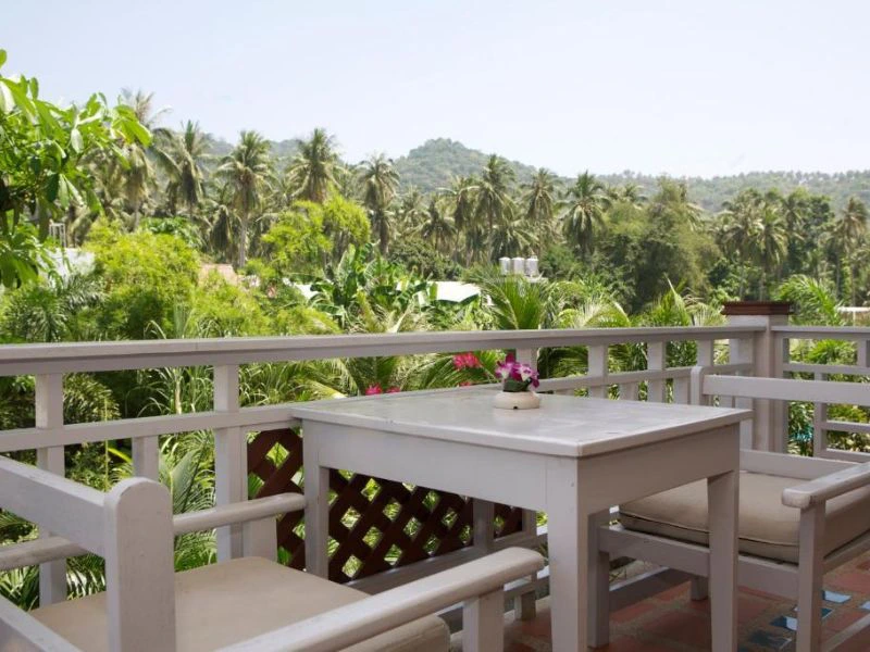 Witte houten tafel met twee stoelen op een balkon, met uitzicht op een groene omgeving vol palmbomen en heuvels op de achtergrond.