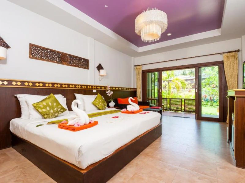Ruime en kleurrijke hotelkamer met een groot bed, sierkussens en handdoeken gevouwen in de vorm van zwanen. De kamer heeft een paarse plafondnis, houten accenten en openslaande deuren naar een groene tuin.