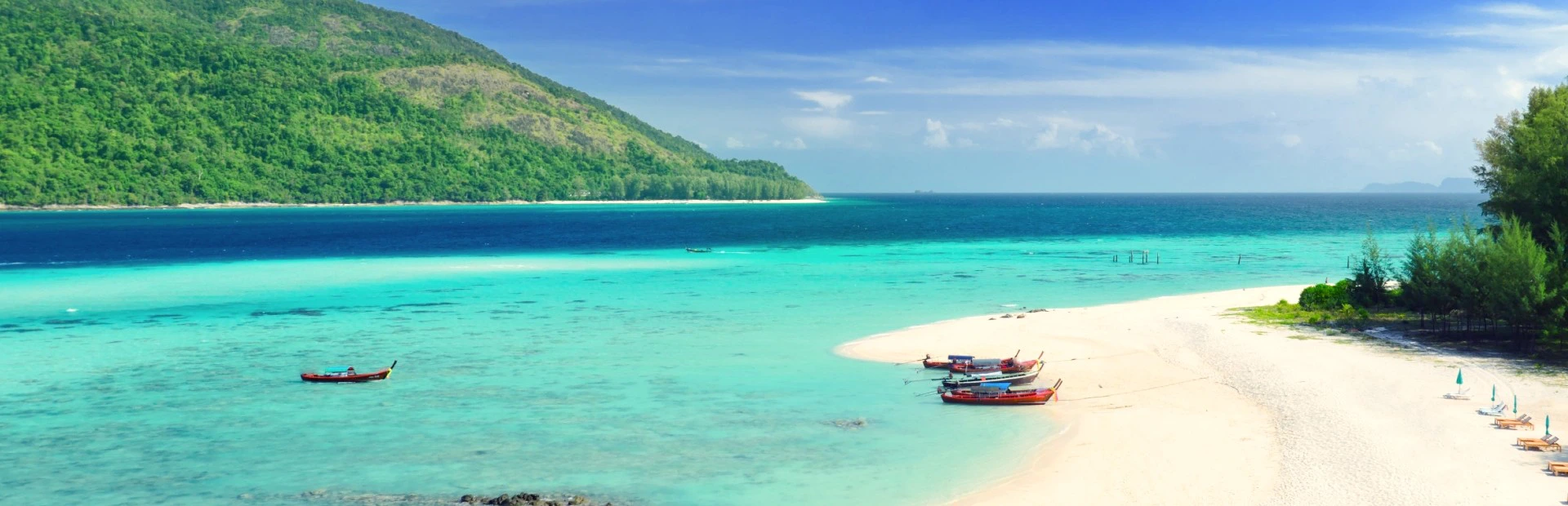Wit zandstrand met turquoise zee, enkele kleurrijke vissersboten in ondiep water en een groen bebost eiland op de achtergrond onder een blauwe lucht.