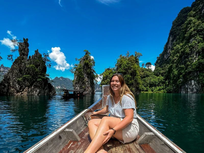 Vrouw zit voorin een houten boot op een meer, omringd door groene kalkstenen rotsformaties en jungle onder een heldere blauwe lucht.