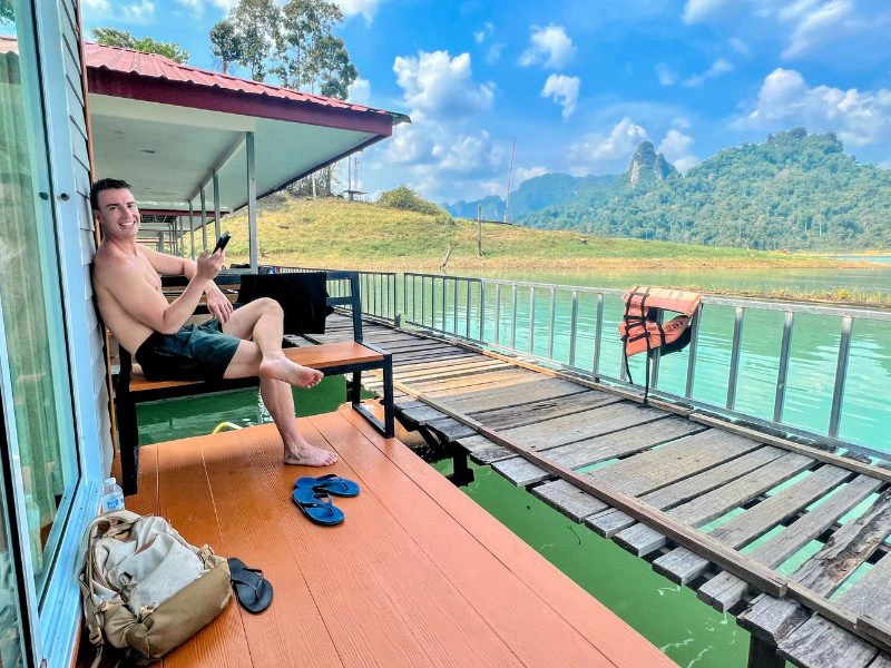 Man ontspant op het terras van een drijvend huisje aan een turquoise meer, met groene heuvels en een blauwe lucht vol wolken op de achtergrond.