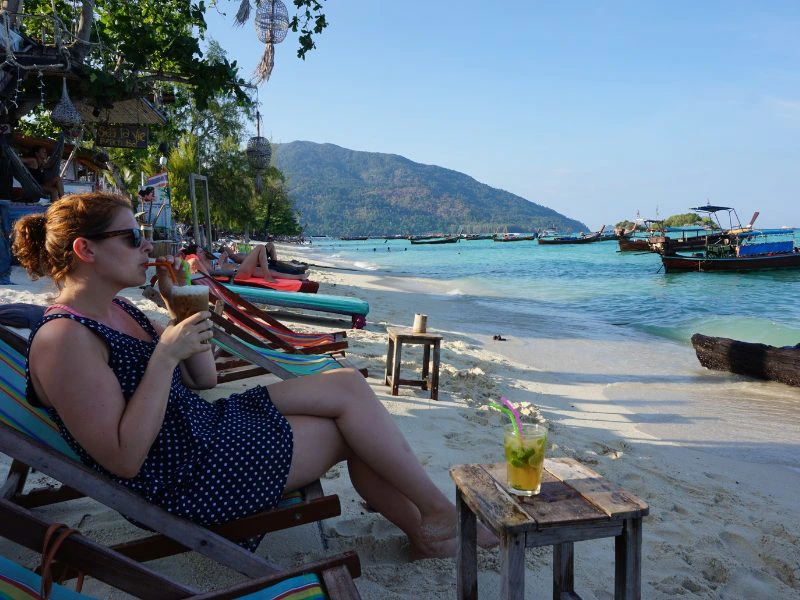 Vrouw met een drankje op een strandstoel. Op de achtergrond longtailboten, een groene heuvel en een blauwe lucht met wat wolken.