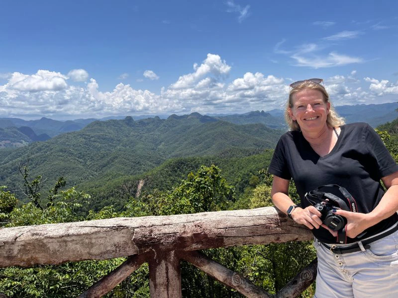 Vrouw met camera leunt lachend op een houten hek, met uitzicht op groene bergtoppen en een helderblauwe lucht vol wolken.