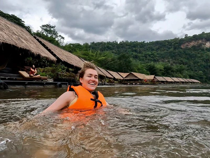 Reizigster met knot op haar hoofd en oranje reddingsvest zwemt in de River Kwai in Thailand. De omgeving heeft groene bergen, grijze lucht en houten huisjes op het water met houten vlonders ervoor.